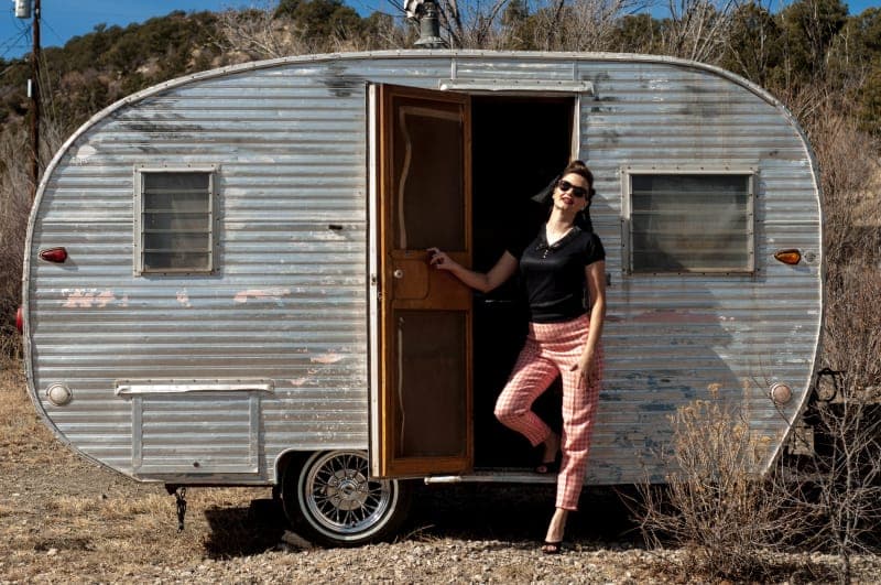 A woman stands in front of a vintage silver trailer with one door open, wearing sunglasses and stylish attire.