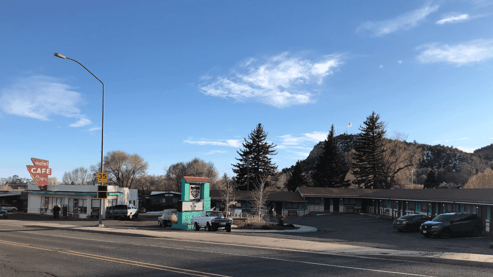 A retro diner sign stands near a motel with teal rooms and a mural, set against a backdrop of trees and hills under a clear blue sky. A retro diner sign stands near a motel with teal rooms and a mural, set against a backdrop of trees and hills under a clear blue sky.