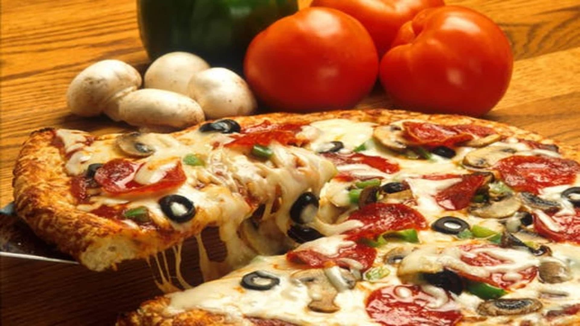 A close-up of a cheese pizza topped with pepperoni, olives, and vegetables, alongside fresh tomatoes, mushrooms, and a green bell pepper.