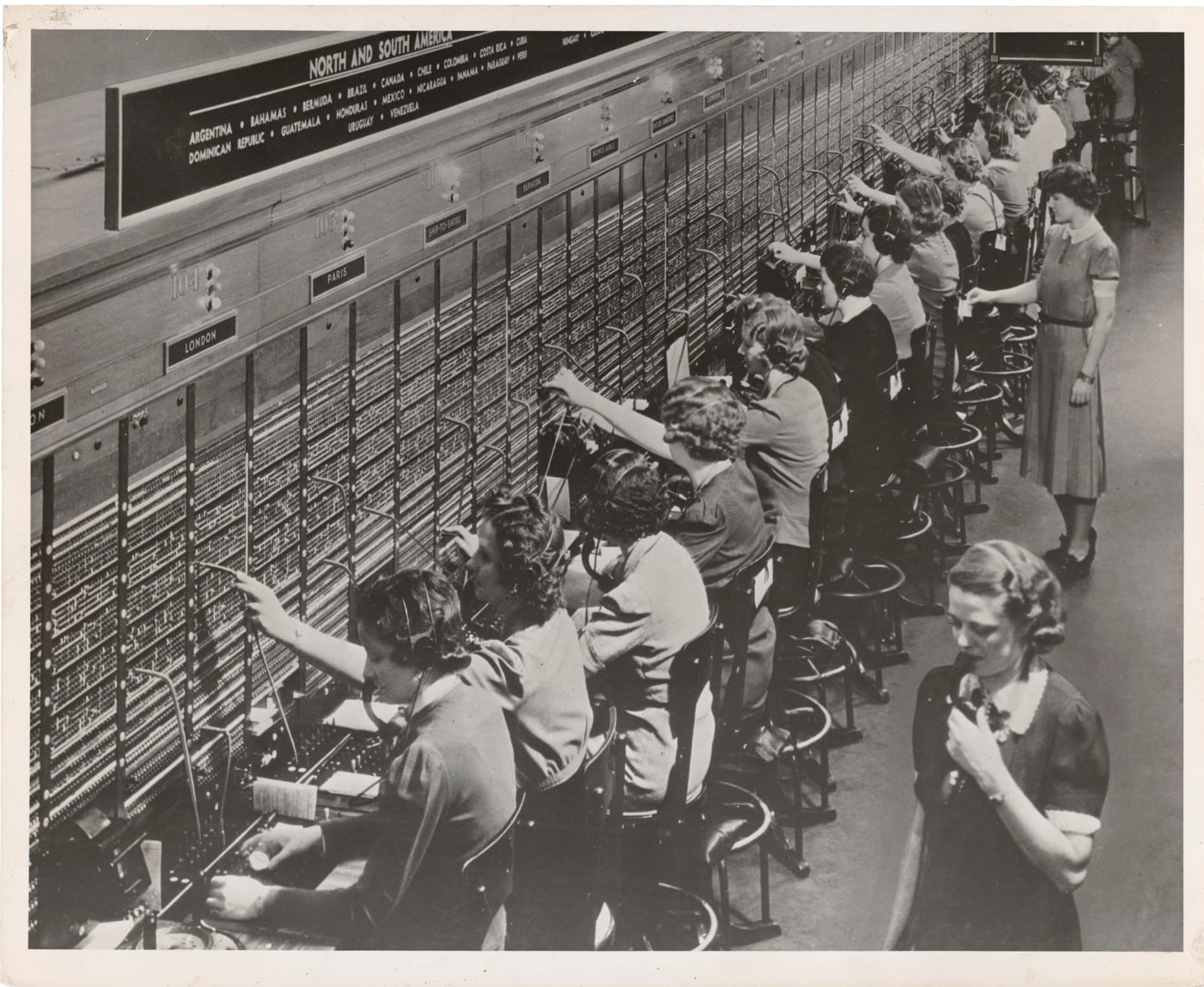 A historical black-and-white photo of multiple telephone operators at switchboards, connecting calls in a busy telecommunications office. A historical black-and-white photo of multiple telephone operators at switchboards, connecting calls in a busy telecommunications office.