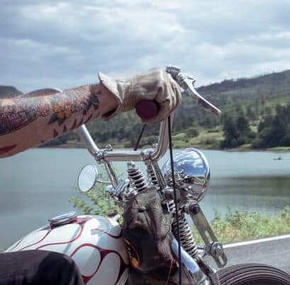 A tattooed hand grips the handlebars of a motorcycle near a scenic lake.