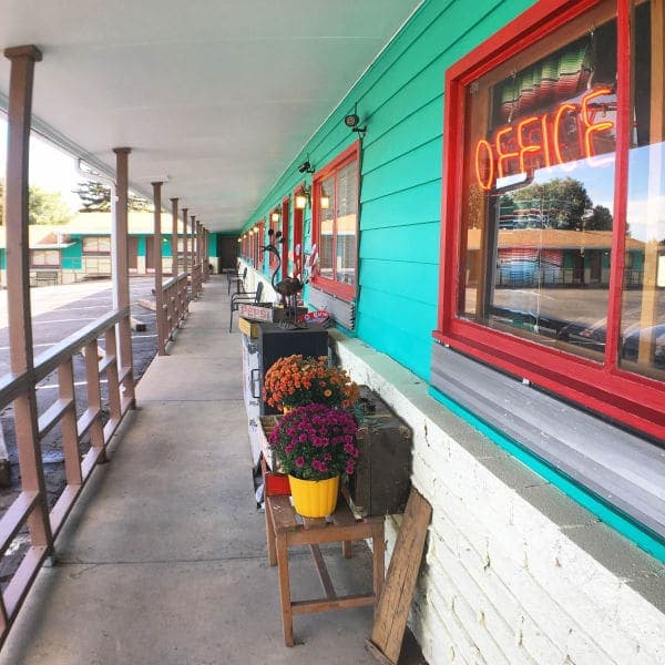 A vibrant motel corridor features potted flowers, a neon "OFFICE" sign, and neatly lined rooms.