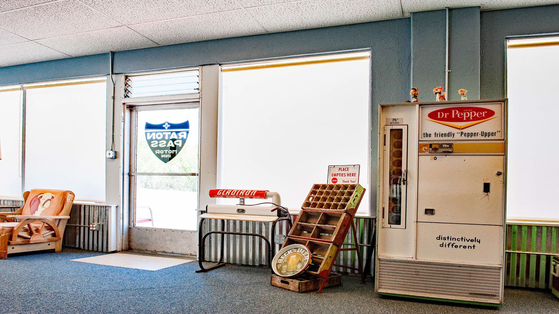 A retro interior featuring a Dr Pepper vending machine, a vintage scale, and an inviting seating area, with large windows covered by blinds.
