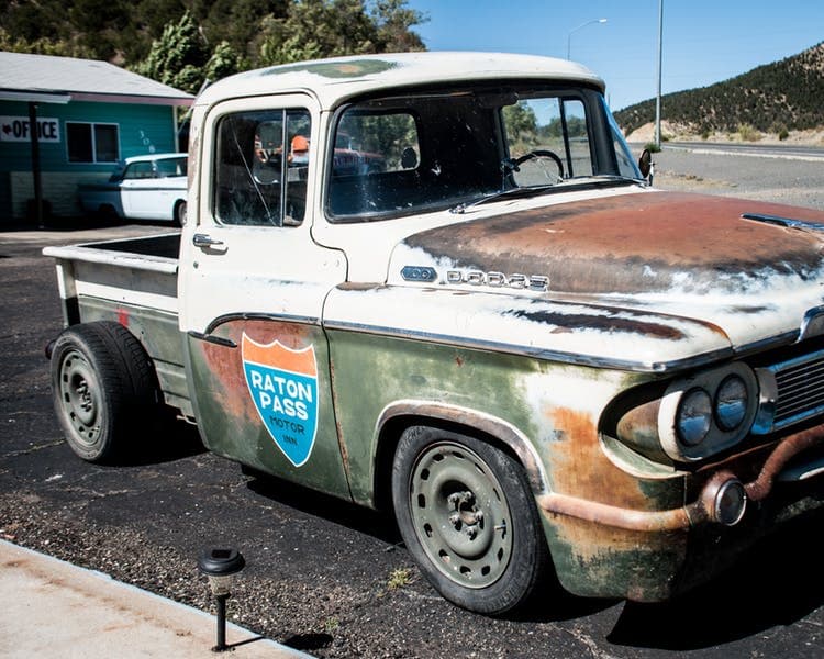 A vintage pickup truck with a weathered green and white exterior featuring a "Raton Pass" logo parked on a rural road.