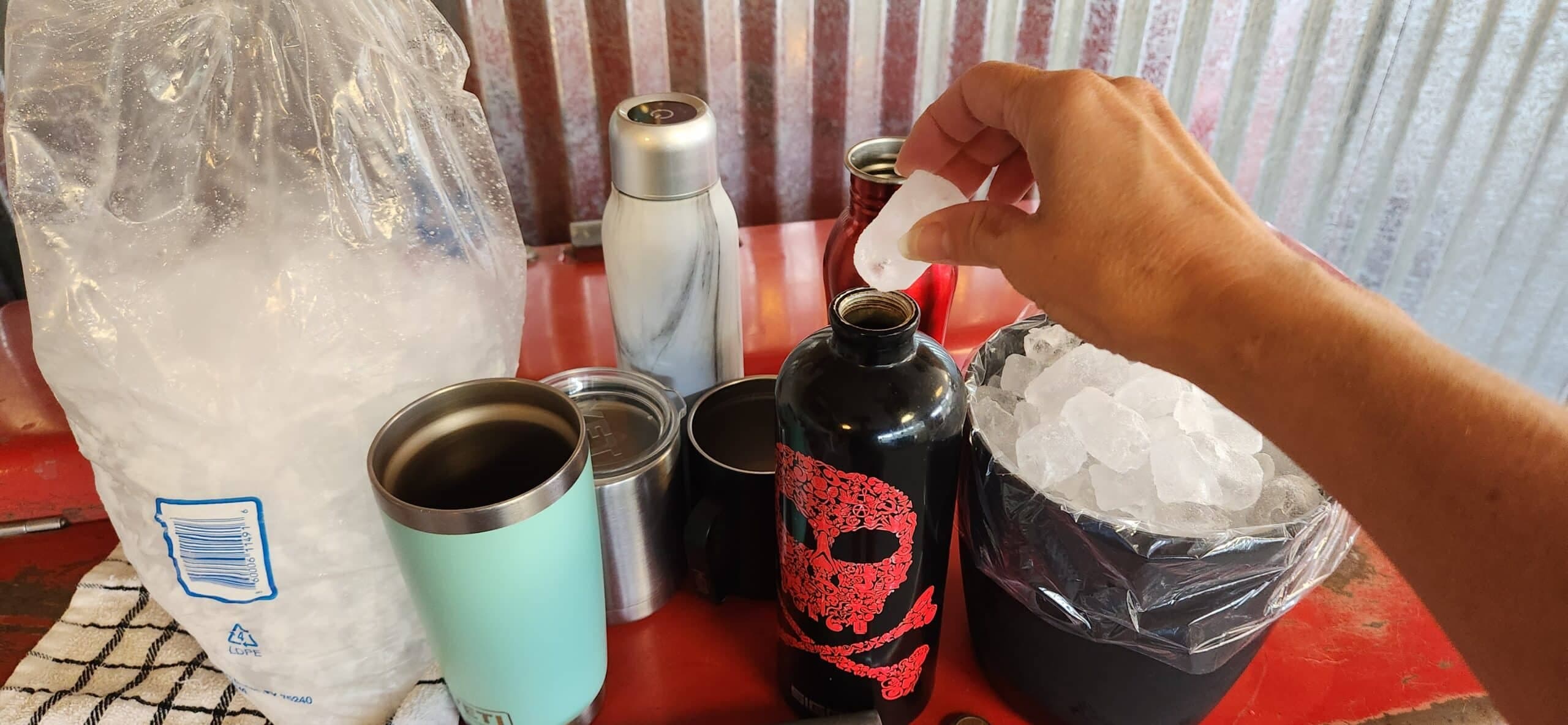 A hand adds ice to a black water bottle surrounded by various containers and bags of ice. A hand adds ice to a black water bottle surrounded by various containers and bags of ice.