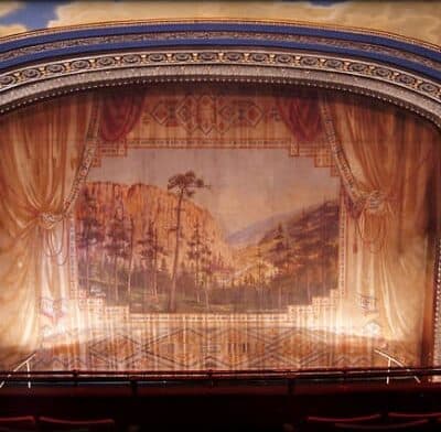 A vintage theater stage with a painted backdrop of mountains and trees, framed by elegant curtains.