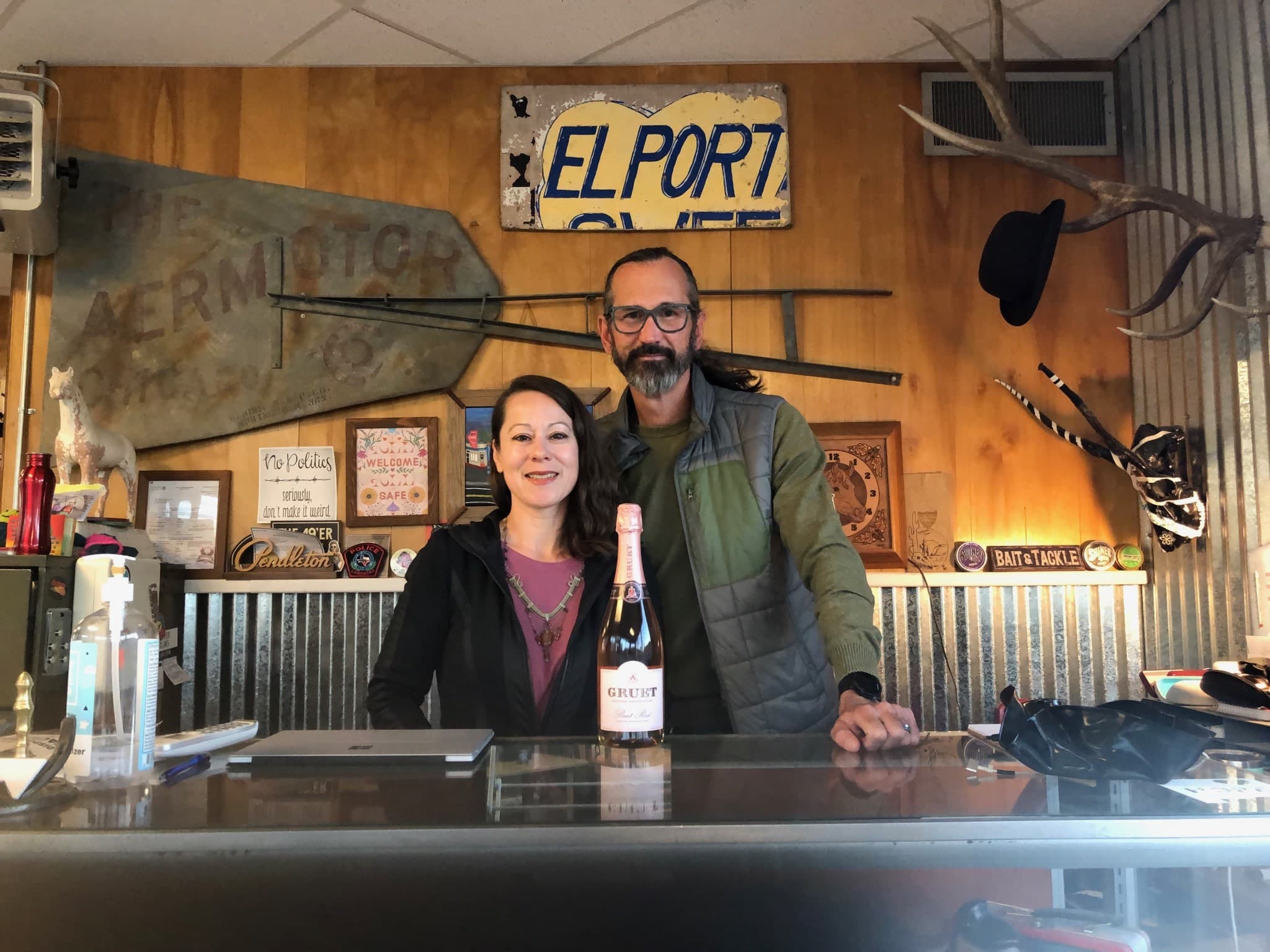 A man and woman stand behind a counter with a bottle of sparkling wine, surrounded by rustic decor and signs. A man and woman stand behind a counter with a bottle of sparkling wine, surrounded by rustic decor and signs.