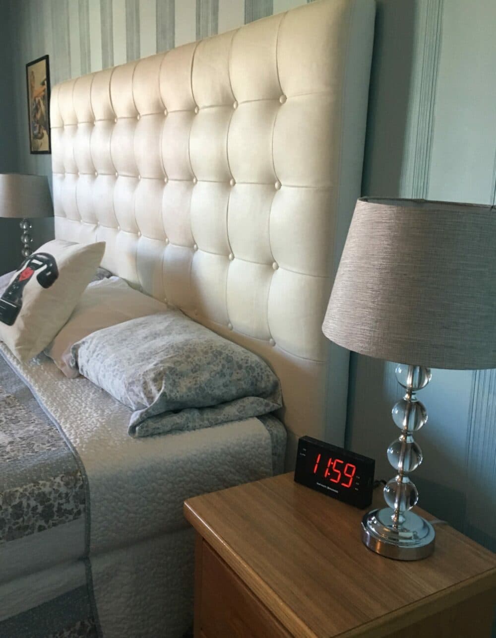 A cozy bedroom with a tufted headboard, decorative pillows, a bedside lamp, and a digital clock displaying 11:59.